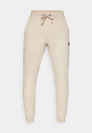 Beige spordipüksid, mis on valmistatud pehmest kangast, millel on paelastringiga vöökoht, küljetaskud ja kitsenevad pahkluupõlved. Lihtne bränding külgedel.