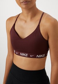 Bordó sportmelltartó V-nyakú kialakítással, állítható vékony pántokkal és az alján kontrasztos fehér Nike logós pánttal. Simább anyagtextúrával.