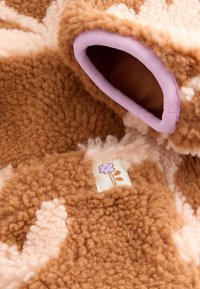 Bruin fleece kledingstuk met een lichtroze accent bij de kraag, zachte textuur en een stoffen label met een bloemmotief.