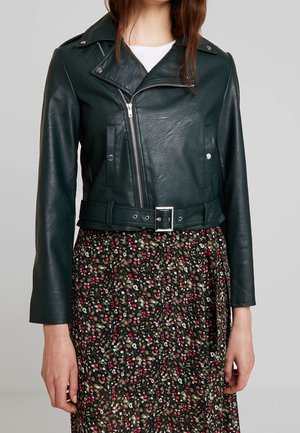 Vrouw die een donker groen leren motorjack draagt met zilveren rits en riem over een rok met een bloemenpatroon in rood, wit en groen.