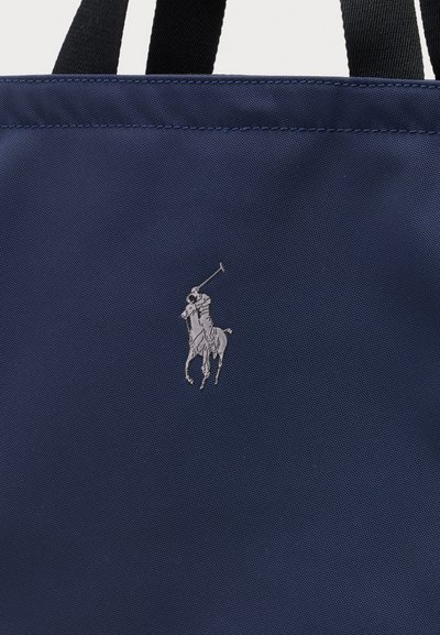 Sac cabas en tissu bleu marine avec deux poignées noires et un logo brodé représentant un cheval et un cavalier tenant un maillet de polo en fil argenté.