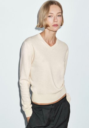 Vrouw met kort blond haar, gekleed in een crèmekleurige trui met V-hals en een zwarte broek, staand tegen een effen lichte achtergrond.