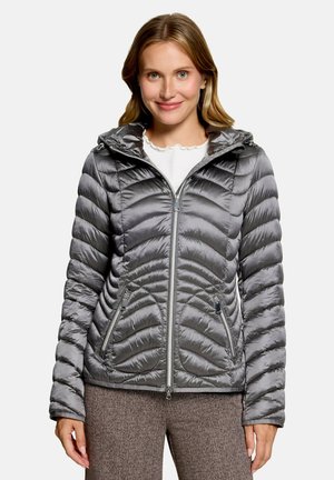 Woman wearing a gray quilted zip-up jacket with a hood and standing against a plain light background.
