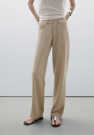 Person wearing beige linen wide-leg pants, white sleeveless top, silver cuff bracelet, and black sandals with metallic decoration standing on gray floor.