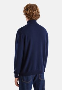 Maglione blu navy con colletto alto, realizzato in tessuto morbido, con polsini e orlo a costine, mostrato da dietro.