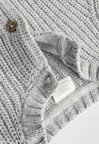 Grau gestrickte Babyjacke mit strukturiertem Muster, ausgestattet mit einem braunen Knopf und einem Etikett mit der Aufschrift "NEXT Baby".