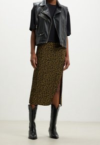Schwarze Leder-Cropped-Weste über einem schwarzen Hemd, kombiniert mit einem braunen Leopardenmuster-Rock mit einem seitlichen Schlitz und schwarzen Cowboy-Stiefeln.