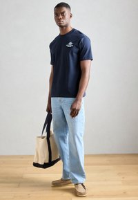 Jūrų laivyno spalvos medvilninis marškinėlis su maža balta grafika, šviesiai mėlyni džinsai, šviesiai rudi slip-on tipo batai ir smėlio spalvos rankinė su tamsiai mėlynomis juostelėmis.