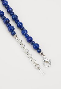 Γαλάζιο κολιέ με χάντρες από γυαλισμένο λάπις λαζούλι, με ασημένια αλυσίδα και κούμπωμα, καθώς και μια μικρή ετικέτα με το εμπορικό σήμα.
