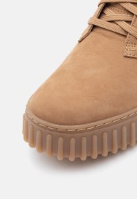Sene i tan suede med en tekstureret overdel, rund tå og en tyk klar gummisål med lodrette ribber. Beige snørebånd fuldender designet.