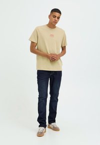 Bež pamučna majica s malim crvenim grafičkim motivom na prsima, kombinirana s tamnoplavim trapericama i sivim tenisicama s bijelim prugama.