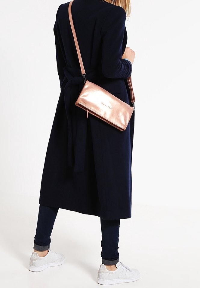 Personne portant un long manteau foncé, un pantalon foncé, des baskets blanches, et un sac bandoulière métallisé rose doré porté sur l'épaule.