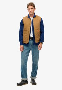 Gilet marron sans manches sur une chemise à manches longues bleue, associ é à un jean bleu clair et des chaussures noires. Design simple, poches pratiques.