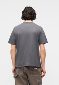 Άτομο με σκούρα κυματιστ ά μαλλιά που φοράει ένα απλό γκρι κοντομάνικο μπλουζάκι και καφέ παντελόνι με μεγάλες τσέπες, με την πλάτη στραμμένη προς τον θεατή.