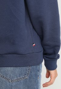Navy blue sweatshirt with ribbed cuffs and hem, featuring a small tricolor logo tag at the side seam. Cotton material, casual fit.