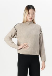 Giovane donna che indossa un maglione beige a collo alto con dettagli in cucitura nera e pantaloni neri, in piedi con una mano sulla vita contro uno sfondo bianco.