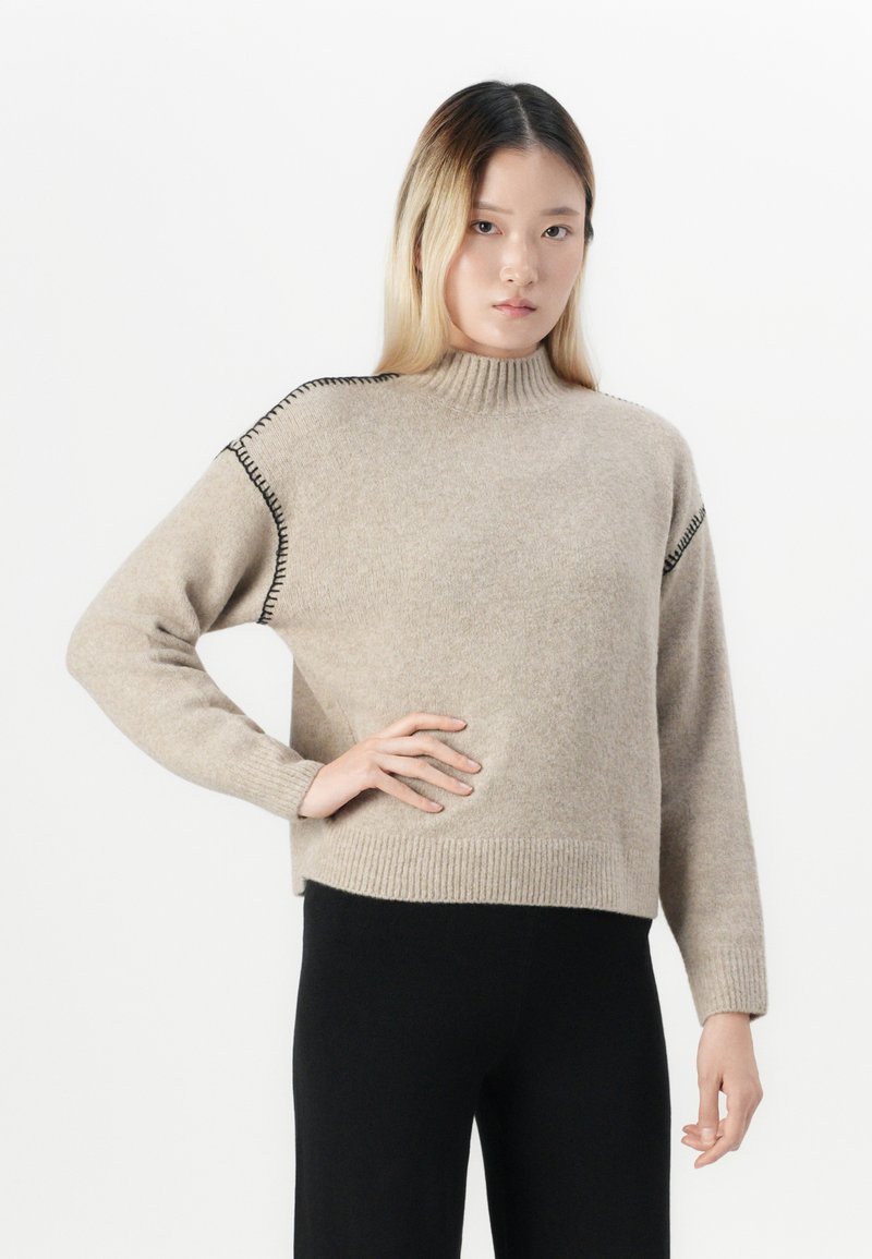 Junge Frau in einem beigen Rollkragenpullover mit schwarzen Stichelementen und schwarzen Hosen, die mit einer Hand auf der Hüfte vor einem weißen Hintergrund steht.