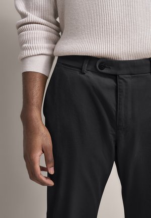 Person trägt einen hellrosa, gerippten Pullover und schwarze Hose, Nahaufnahme der Hand- und Taillenpartie.