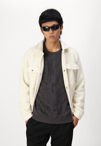 Jachetă albă din fleece cu capse peste un pulover texturat închis. Modelul poartă ochelari de soare negri și pantaloni negri, pozând pe un fundal alb.