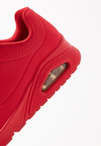 Red athletic sneaker with a smooth, textured upper, perforated details, cushioning air unit, and a rubber outsole featuring traction grooves.
