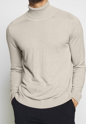 Homme portant un pull à col roulé beige avec des poignets et un ourlet côtelés, associé à un pantalon noir à fines rayures, les mains dans les poches.