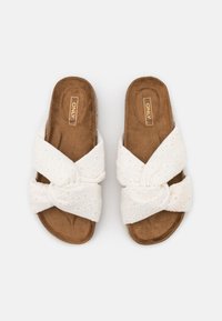 Sandales à glissière blanches avec des brides croisées ornées de motifs en œillets et un détail de nœud. Semelles intérieures souples couleur marron avec une finition texturée.
