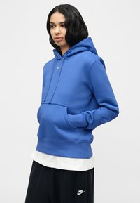 Άτομο που φοράει μπλε φούτερ με κουκούλα Nike με τα χέρια στην μπροστινή τσέπη και μαύρο παντελόνι Nike, στέκεται μπροστά σε απλό ανοιχτό φόντο.