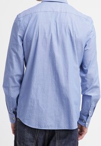 Sininen pitkähihainen paita, jossa on hienovarainen pistekuvio, napilliset kalvosimet, klassinen kaulus ja kaareva helma. Valmistettu teksturoidusta kankaasta.