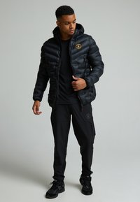 Svart pufferjacka med huva, dragkedja fram och röda detaljer. I kombination med svarta cargo-byxor och svarta sneakers.