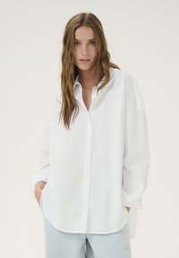White oversized button-up shirt made of a smooth fabric. Features a classic collar, long sleeves, and a rounded hem. Simple design.