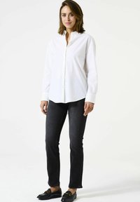 Chemise blanche à boutons avec manches longues, associée à un jean foncé. La tenue présente une coupe classique et des mocassins noirs contrastants.