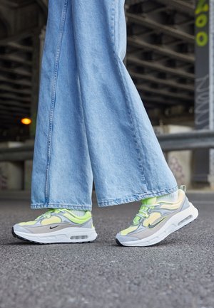 Persoon die lichtblauwe wijde jeans en neon groene en grijze Nike sneakers draagt, staand op asfalt onder een brug.