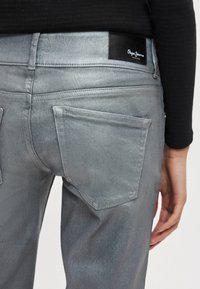 Hallid teksariided kitsama lõikega, millel on tagataskud, peenike õmbluste detailid ja vöökoha juures must nahast märk logo kujutisega.