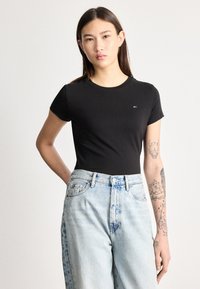 Ung person med langt hår iført en sort tætsiddende t-shirt og lyseblå jeans med høj talje, der viser tatoveringer på venstre arm.