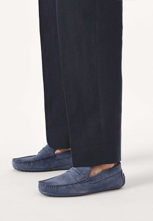 Mocassins en daim bleu marine portés avec un pantalon noir, montrant un style décontracté slip-on avec couture mocassin et semelles en caoutchouc.