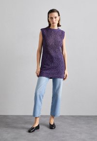 Violeta bezpiedurkņu adīta tunika ar faktūru, sapārota ar gaiši ziliem taisnajiem biksēm un melniem slip-on apaviem.