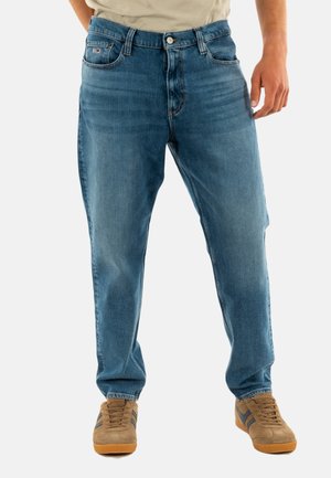 Vaqueros de mezclilla azul claro con corte recto, cinco bolsillos y cierre de botón. Combinados con zapatillas marrones con rayas oscuras.