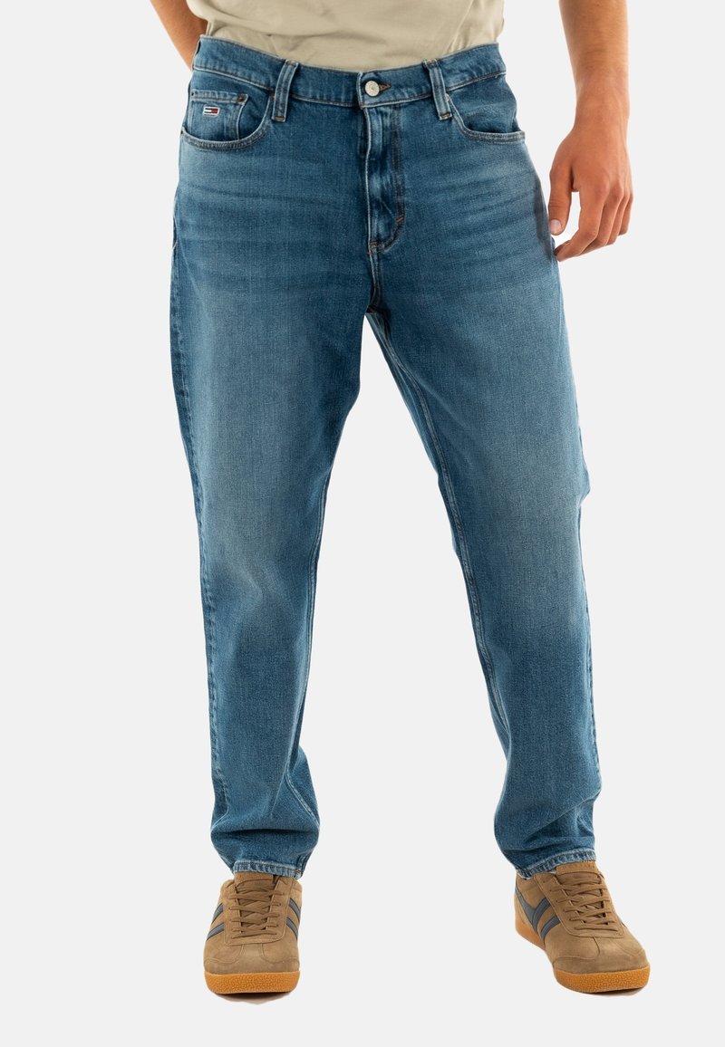 Vaqueros de mezclilla azul claro con corte recto, cinco bolsillos y cierre de botón. Combinados con zapatillas marrones con rayas oscuras.