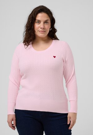 Femme portant un pull en maille torsadée rose clair avec un petit emblème en forme de cœur rouge, associé à un jean bleu foncé, debout devant un fond gris.