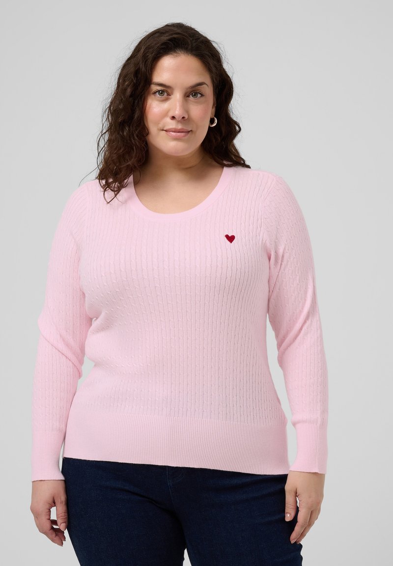 Femme portant un pull en maille torsadée rose clair avec un petit emblème en forme de cœur rouge, associé à un jean bleu foncé, debout devant un fond gris.