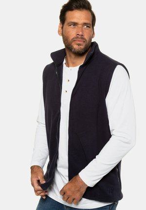 Navy fleece vest with zip closure, high collar, and side pockets, worn over a white long-sleeve shirt, featuring a casual fit and smooth texture.