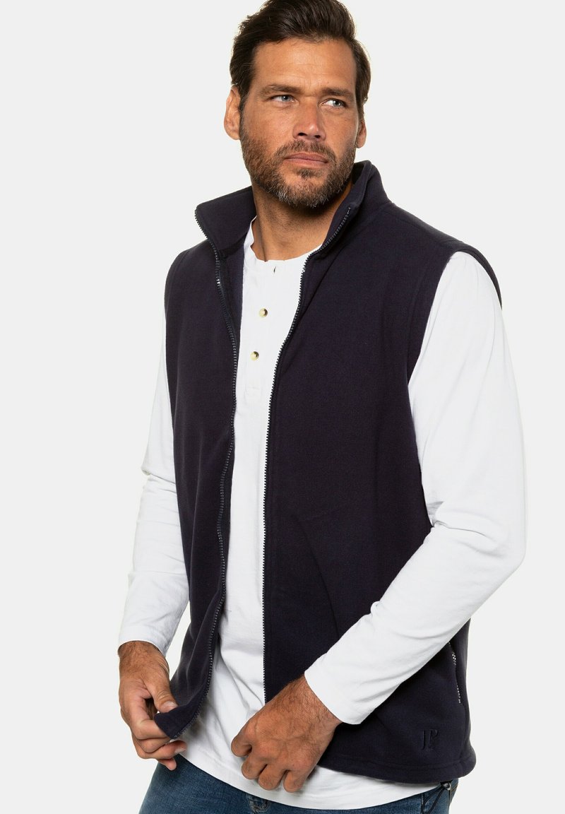 Gilet in fleece blu navy con chiusura a zip, colletto alto e tasche laterali, indossato sopra una camicia bianca a maniche lunghe, con una vestibilità casual e una texture liscia.