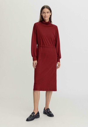 Rood, lang mouwend dress met een col, elastische tailleband en midi-lengte. Gepaard met zwarte loafers, gladde stof textuur.