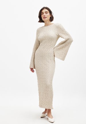 Cream, long-sleeve, fitted dress with textured fabric, round neckline, and flared sleeves, reaching mid-calf. Paired with light-colored shoes.