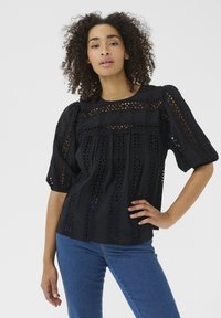 Sort bluse med øjelet detaljer, buet halsudskæring, korte puffærmer og lagdelt tekstur. Parret med blå jeans for kontrast.
