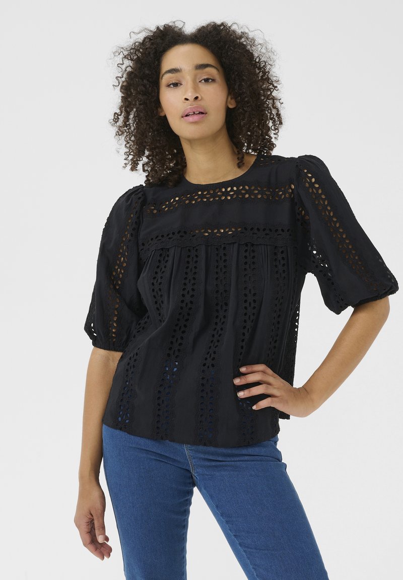 Sort bluse med øjelet detaljer, buet halsudskæring, korte puffærmer og lagdelt tekstur. Parret med blå jeans for kontrast.