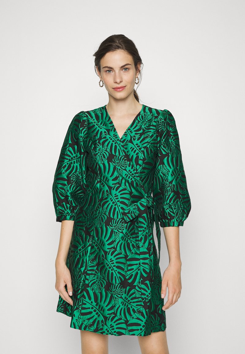 Robe portefeuille verte avec un motif de feuilles noires, dotée de manches bouffantes et d'une fermeture à nouer à la taille, fabriquée dans un tissu lisse et brillant.