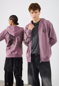 Ανοιχτό μωβ φούτερ με φερμουάρ και γραφικό σχέδιο; γκρι μπλούζα από κάτω; μαύρα φθαρμένα παντελόνια; φθαρμένη εμφάνιση με ξέφτια.