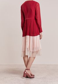 Vestido midi plisado rojo con una capa superior translúcida, que transita hacia una falda de color rosa más claro. Presenta mangas largas y zapatos de tacón rosa.