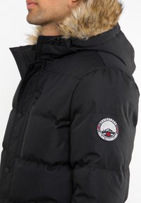 Sort pufferjakke med en aftagelig hætte foret med pels, quiltet tekstur, lynlåslommer og et logo-patch på venstre ærme.
